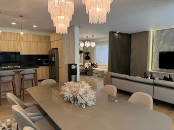 Contemporary open-plan apartment interior: glossy taupe dining table with dried floral centerpiece, upholstered dining chairs, wood-grain kitchen with breakfast bar and stools, cascading chandeliers, neutral sectional sofa and marble-accent TV wall with warm ambient lighting.