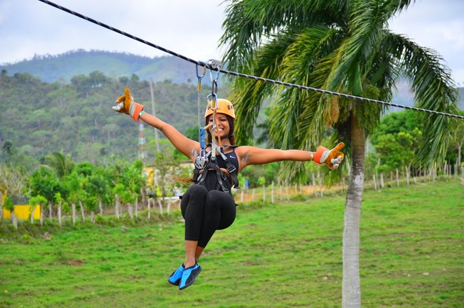 Person ziplining with arms outstretched, wearing helmet and harness, gliding past palm trees over green fields with a mountain backdrop — tropical outdoor adventure.