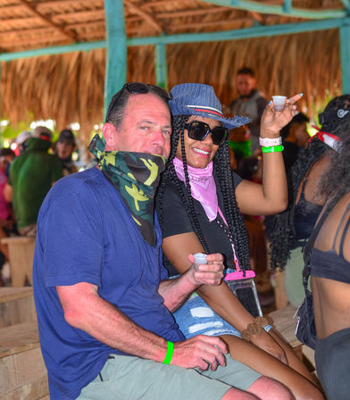 Two friends at a tropical thatched-roof beach bar raising shot cups, smiling and wearing bandanas, sunglasses, and casual summer clothes.