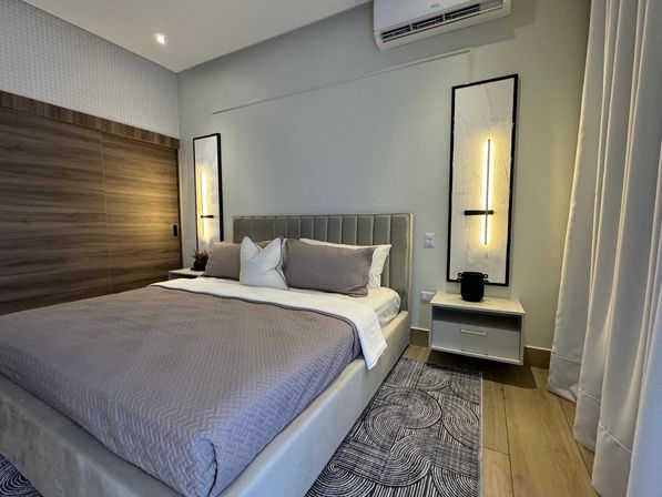 Modern bedroom interior with upholstered bed and gray quilt, decorative pillows, floating nightstands with vertical LED wall sconces, wood sliding closet, patterned area rug, curtains and wall-mounted AC.