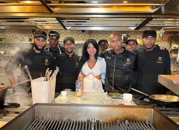 Smiling guest in a white dress posing with a team of professional chefs in black uniforms in a busy restaurant open kitchen with stainless-steel stovetops, utensils, pans and patterned tile backsplash.
