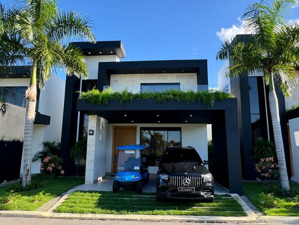 Modern two-story tropical house with black geometric façade, balcony planters, palm trees and manicured lawn; driveway with a blue golf cart and black luxury SUV under a clear blue sky