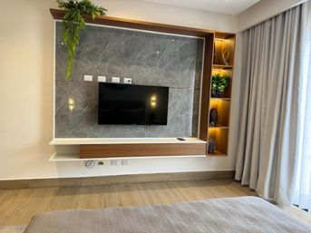 Modern bedroom TV feature wall with gray marble backdrop, wall-mounted flat-screen above a wood floating console, built-in illuminated shelving with greenery, light-gray floor-to-ceiling curtains and wood-plank flooring.
