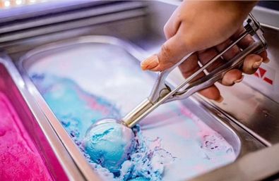 Bright blue-and-pink cotton-candy ice cream being scooped from a stainless pan at an ice cream shop counter.
