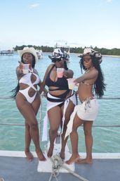 Three women in swimsuits and cowgirl hats toasting with drinks on a boat deck during a tropical boat party, turquoise water and palm‑fringed island in the background.