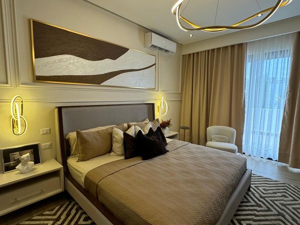 Contemporary neutral master bedroom with quilted taupe bedding, layered pillows, glowing wall sconces, abstract wall art and floor-to-ceiling curtains over a balcony door.