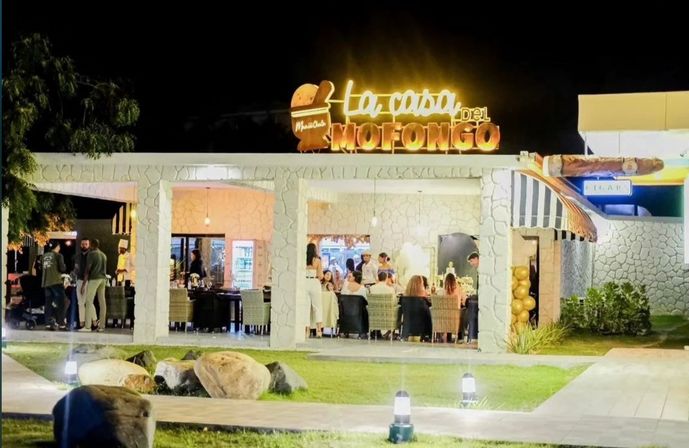 Nighttime outdoor Puerto Rican mofongo restaurant with stone-faced open-air terrace, warm neon sign, diners at tables, grassy courtyard with decorative rocks and path lights.