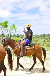 Person in a yellow helmet and sunglasses taking a selfie while riding a brown horse on a sunny tropical trail with palm trees and blue sky
