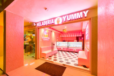 Neon-pink ice cream parlor storefront with glass doors, pink banquette seating, striped awning, illuminated gelato display freezers and black-and-white hex tile floor