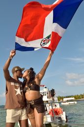 Couple kissing on a boat holding a large Dominican Republic flag overhead, with turquoise Caribbean water, other boats and a palm-lined shore under a clear blue sky
