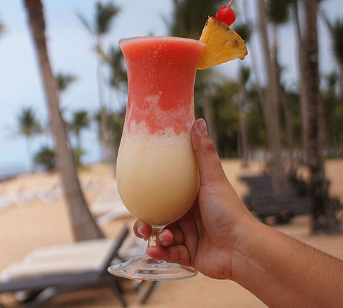 Hand holding a layered frozen tropical cocktail (pineapple yellow and strawberry red) garnished with a pineapple slice and cherry, with palm trees and beach lounge chairs blurred in the background.