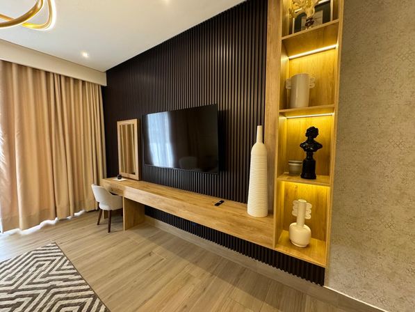 Modern living room interior with wall-mounted flat-screen TV on a black slatted accent wall, long floating wooden console, built-in illuminated shelves with decorative vases and bust, beige curtains, small chair and patterned rug on wood flooring