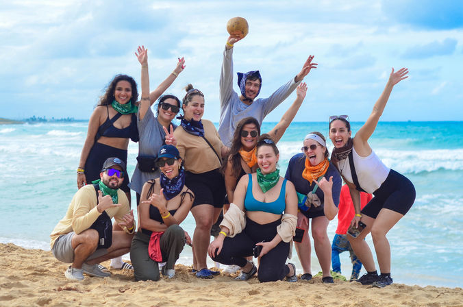 Energetic group of friends posing on a sandy tropical beach with turquoise waves, wearing colorful bandanas and beachwear, one person holding a coconut and everyone cheering