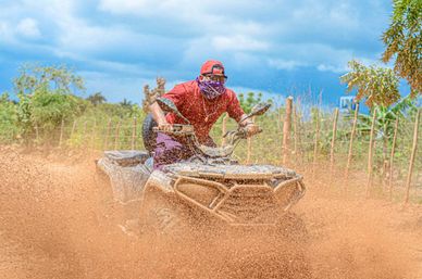 ATV rider in red shirt and cap with a purple bandana powering through a muddy off-road countryside trail, mud spraying under a bright blue sky