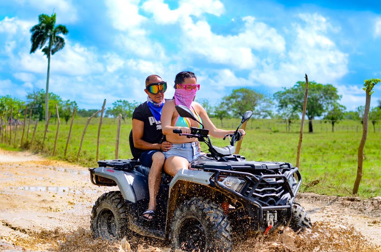 Two riders on an ATV splashing through a muddy rural trail under a bright blue sky, wearing goggles and bandanas with a palm tree and green pasture in the background — off-road tropical adventure.