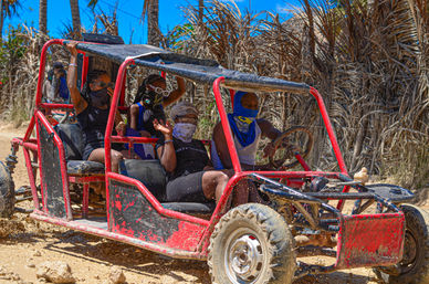 Four masked passengers in sunglasses ride a red off-road dune buggy, one waving, as mud and dust spray on a sunny tropical dirt trail lined with palm trees and dried foliage.