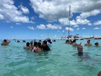 Vacationers wading in clear turquoise shallow water near anchored catamarans and sailboats under a bright blue sky dotted with fluffy clouds