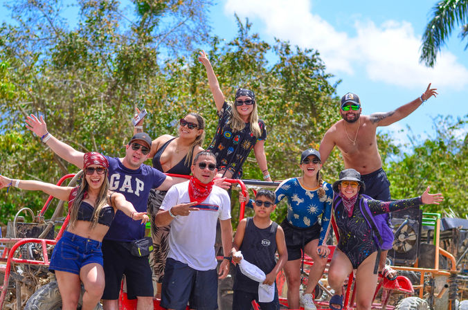 Smiling group of friends in swimwear, sunglasses and bandanas posing on a red off-road dune buggy during a sunny tropical dune-buggy tour with palm trees and blue sky.
