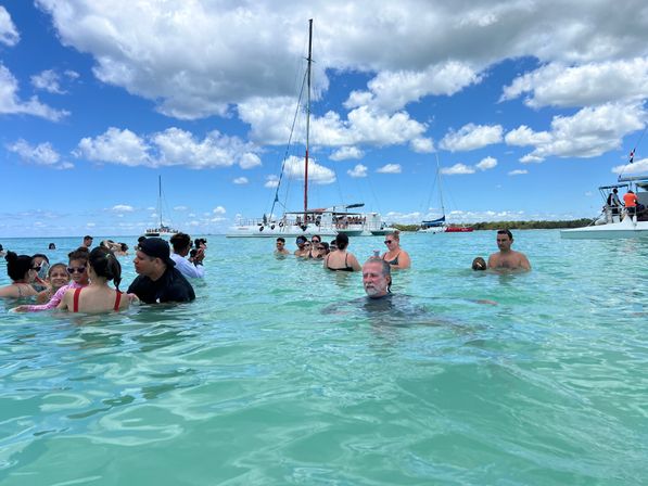 People enjoying a tropical beach day, wading in clear turquoise water with anchored catamarans and sailboats under a bright blue sky with fluffy clouds