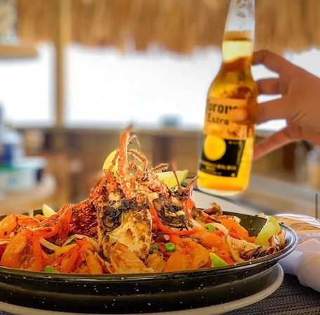 Tropical beachside seafood skillet topped with grilled lobster, rice, peas and peppers, lime wedges and a hand holding a beer