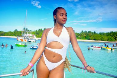 Woman in a white cut-out swimsuit posing on a boat railing over turquoise tropical waters, with swimmers, colorful speedboats, a palm-lined island shore and bright blue sky — sunny vacation scene.