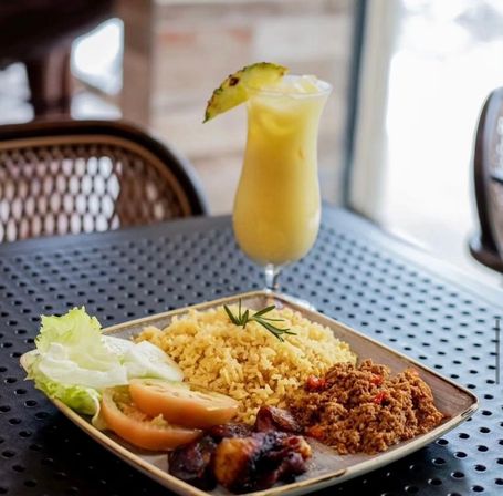 Colorful tropical meal: plate of seasoned yellow rice with spiced minced meat, fried plantains and tomato-lettuce salad, paired with a pineapple-topped tropical juice on a perforated outdoor café table.