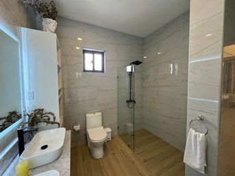Contemporary bathroom interior with marble-look tiled walls, wood-look plank floor, glass walk-in shower with black fixtures, white toilet, vessel sink on a marble vanity and towel ring.