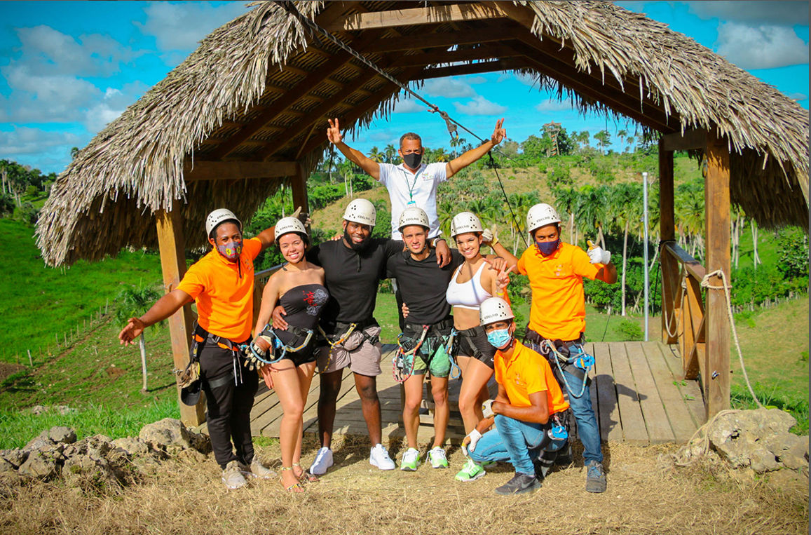 Cheerful group of adults in helmets and harnesses on a thatched zipline platform with guides in orange shirts, set against tropical palm-covered hills and blue sky — outdoor zipline adventure