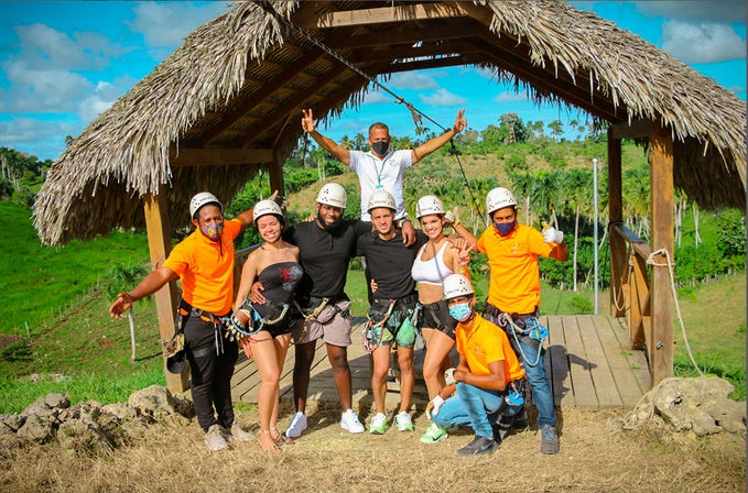 Cheerful group of adults in helmets and harnesses on a thatched zipline platform with guides in orange shirts, set against tropical palm-covered hills and blue sky — outdoor zipline adventure