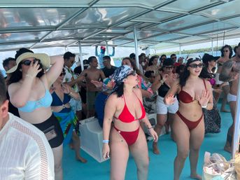 Crowd of people dancing and socializing on a covered catamaran boat party in sunny tropical turquoise waters, many wearing bikinis and beachwear.