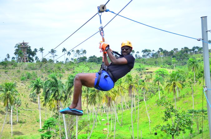Person wearing an orange helmet and sunglasses ziplining over a lush, palm‑studded tropical hillside — outdoor adventure zipline in a green landscape.