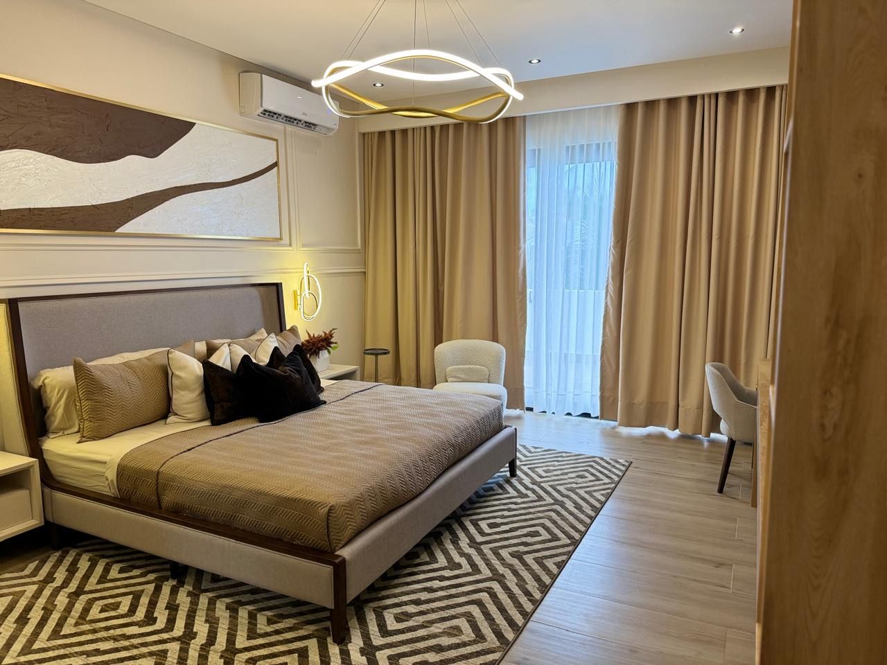 Contemporary beige bedroom interior — upholstered king bed with layered pillows, geometric area rug, modern circular pendant light, cozy armchairs and floor-to-ceiling curtains.