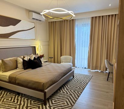 Contemporary beige bedroom interior — upholstered king bed with layered pillows, geometric area rug, modern circular pendant light, cozy armchairs and floor-to-ceiling curtains.