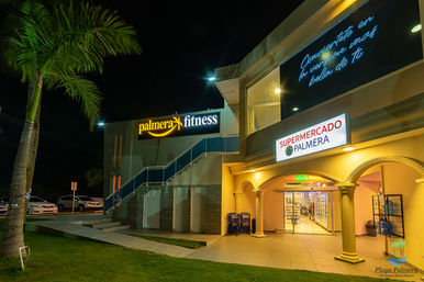 Night scene at a tropical beach resort complex featuring an illuminated fitness center and supermarket entrance, palm tree, tiled walkway and neon signage.