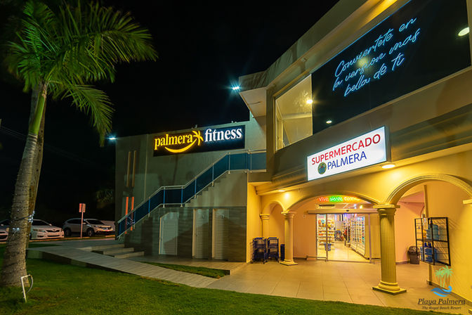 Night scene at a tropical beach resort complex featuring an illuminated fitness center and supermarket entrance, palm tree, tiled walkway and neon signage.