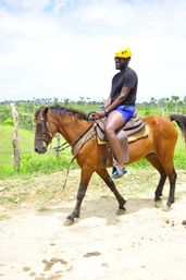 Smiling rider in a yellow helmet and blue shorts on a brown horse trotting along a dirt trail in a lush tropical countryside with palm trees