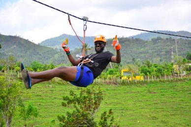 Adventurer in helmet, sunglasses and harness gives thumbs-up while ziplining over lush green hills and distant mountains — outdoor zipline adventure in countryside.