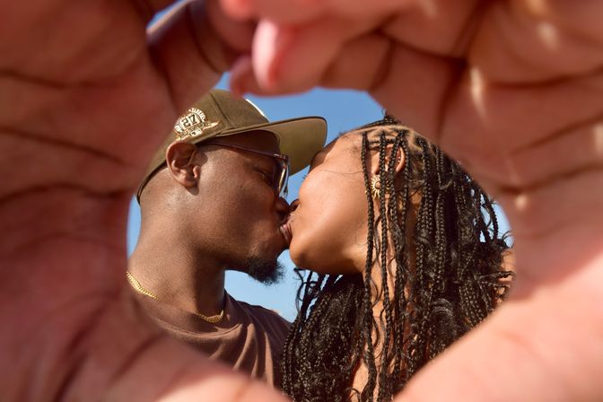 Romantic couple kissing outdoors on a sunny day, framed through hands shaped into a heart; woman with long braids and man wearing a cap and glasses against a clear blue sky.