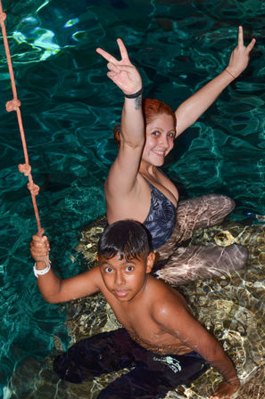 Smiling woman and boy in a turquoise resort swimming pool, boy holding a rope while the woman flashes peace signs — playful vacation swim scene.
