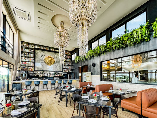 Luxury restaurant interior with soaring ceilings, two grand crystal chandeliers, marble-backed bar stocked with bottles and gold lion wall art, lush planter ledge, orange banquette seating and neatly set dining tables by floor-to-ceiling windows.