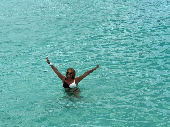 Woman in black-and-white bikini and sunglasses, waist-deep in clear turquoise tropical water with arms raised in a joyful vacation pose