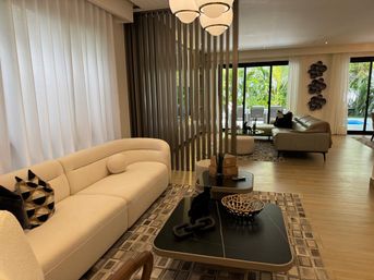 Sunlit modern open-plan living room with a curved cream sofa, geometric throw pillows, black marble coffee table with decorative bowl and chain sculpture, vertical slatted room divider, sheer floor-to-ceiling curtains and sliding glass doors revealing a lush tropical garden and pool.