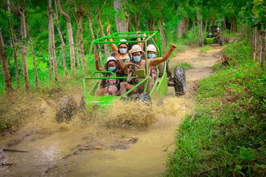 Group of people in helmets and face masks riding a lime-green off-road buggy through a muddy tropical forest trail, splashing water and cheering in an adventurous off-road tour.