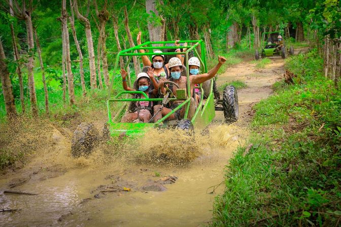 Group of people in helmets and face masks riding a lime-green off-road buggy through a muddy tropical forest trail, splashing water and cheering in an adventurous off-road tour.