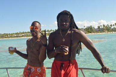 Two shirtless men on a boat holding plastic cups of beer — one wearing mirrored sunglasses and flashing a peace sign, the other with long dreadlocks — crystal-clear turquoise water and a palm-lined tropical beach under a sunny blue sky.