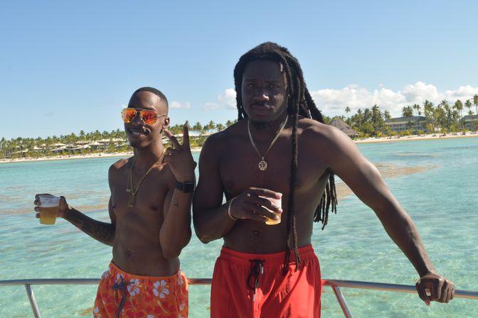 Two shirtless men on a boat holding plastic cups of beer — one wearing mirrored sunglasses and flashing a peace sign, the other with long dreadlocks — crystal-clear turquoise water and a palm-lined tropical beach under a sunny blue sky.