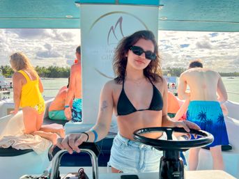 Woman in black bikini top and denim shorts steering a catamaran on a sunny tropical cruise, passengers lounging on deck with palm-lined shore and turquoise water under a partly cloudy sky.