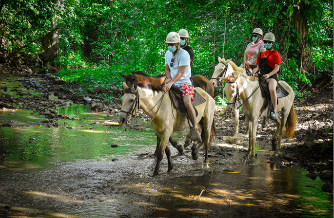 Four helmeted riders wearing face masks on horseback crossing a shallow tropical forest stream beneath a dense green canopy