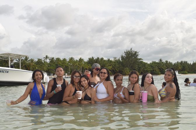 Smiling group of friends in swimsuits standing waist-deep in clear shallow tropical water near a palm-lined beach and anchored boat, holding drinks under a cloudy sky.