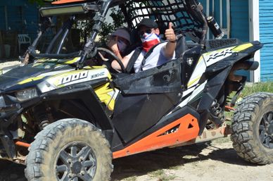 Off-road side-by-side UTV with muddy tires and two masked passengers giving a thumbs-up in a sunny outdoor scene by a blue wooden building.